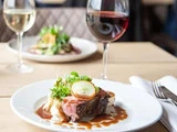 En anrettet bøfret med sovs, kartoffelmos og grøntsager, med et glas rødvin i baggrunden på et træbord.