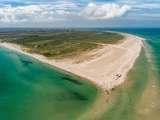 Luftfoto af en sandhalvø omgivet af turkisblåt vand med nogle få mennesker og køretøjer langs stranden. Grøn vegetation dækker området inde i landet.