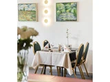 Et lille restaurantbord til fire med hvid dug, sirligt foldede servietter og en enkelt blomst i en vase. To landskabsmalerier og bløde væglamper pynter på den hvide væg.