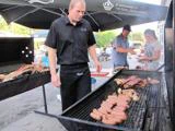 En mand griller pølser og kød udendørs, mens andre tilbereder mad i nærheden under store parasoller ved et grillarrangement.