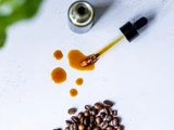 En dråbetæller med mørk væske, tre dråber, en flaske og ristede kaffebønner på en hvid flade.