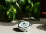 En lille rund dåse mærket "multi balm" med et lille bogstav "g" på låget, placeret på en lys flade med grønne bladplanter i baggrunden.