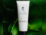 En tube med Grums raw coffee hand scrub and wash står oprejst på grønne blade. Den hvide tube har sorte prikker og minimalistisk tekst, der fremhæver den økologiske kaffegrums og den danske oprindelse