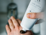 En person klemmer en mørk scrub fra en hvid tube mærket raw coffee hand scrub ud på håndryggen.