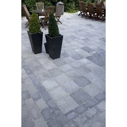 En gårdhave med grå stenbelægning, to høje rektangulære krukker med små stedsegrønne buske og træborde og -stole i baggrunden.