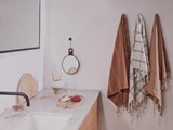 Minimalistisk badeværelse med en hvid bordplade, et lille rundt spejl, pyntegenstande og tre håndklæder - to brune og et hvidt med striber - der hænger på væggen.