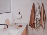 Et moderne badeværelse med marmorbordplade, sort vandhane, rundt spejl og tre håndklæder på væggen i brune og hvide toner. Minimalistiske indretningsgenstande står på køkkenbordet.