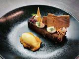 En platedessert består af et stykke kage toppet med creme, pynt og sprøde skår, serveret med en kugle lys is på frugtpuré, alt sammen på en sort tallerken.