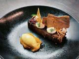 En sort tallerken med et stykke kage toppet med creme, krydderurter og tynde, sprøde vafler, serveret med en kugle gul sorbet på en frugtpuré.