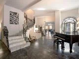 Elegant foyer med marmortrappe, udsmykket sort gelænder, sort flygel, stort vindue og moderne kunst på væggen. Rummet har polerede stengulve og varm belysning.