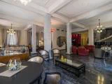 Elegant hotellounge med marmorgulv, runde borde, polstrede stole, glasindretning, lysekroner, røde lænestole og en trappe med udsmykkede gelændere i baggrunden.