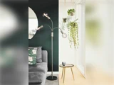 En moderne stue med en grå sofa, en sort gulvlampe, hængende planter, et rundt spejl og et lille rundt sidebord ved et lyst vindue.