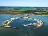 Luftfoto af en trekantet marina med dokker og både, omgivet af land, grønne marker og vand, under en klar blå himmel.