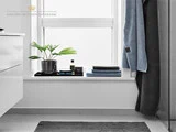 Moderne badeværelse med vindue, potteplante, toiletartikler og pænt foldede blå håndklæder i vindueskarmen. Et mørkt håndklæde hænger i nærheden; et gråt tæppe ligger på det hvide gulv.