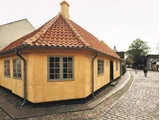 Traditionelt gult hus med rødt tegltag og grå vinduer på en brostensbelagt gade, med et par mennesker gående i baggrunden.