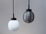 To ribbede pendellamper hænger side om side mod en almindelig væg; den ene lampe er hvid, og den anden er røggrå. Begge har sorte ledninger og cylindriske, sorte beslag.