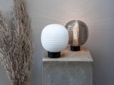 To runde, riflede bordlamper - en hvid og en røggrå - står på en betonsokkel ved siden af tørret pampasgræs på en lysegrå væg.