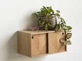 En lille væghylde af træ med skydelåger, der rummer en glasvase med en bladplante og en lille lyserød pyntegenstand på toppen.