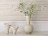 Hvide vilde blomster i en rund beige vase ved siden af to mindre beige keramiske vaser på en lys overflade, med en minimalistisk baggrund.
