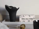 En sort vase og en æske med påskriften »Nabos Lilla Telåda« står på et hvidt fad ved siden af en gylden tefilter på en lys overflade.