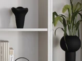 En sort vase på en hvid hylde, bøger stablet nedenunder og en sort, rund vase med grønt løv til højre.