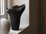 En sort vase med bølgede kanter og et snoet mønster står på en vindueskarm ved siden af et rundt, hvidt objekt, der er blidt oplyst af naturligt lys.