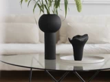 På et moderne sofabord af glas står en høj, sort vase med grønne planter og et mindre, skulpturelt, sort objekt på et hvidt fad, med en beige sofa i baggrunden.