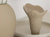 En beige, abstrakt keramisk vase med et bølget, snoet mønster står på et rundt fad ved siden af et glat, rundt objekt i en minimalistisk opstilling.
