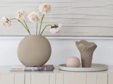 Rund vase med lyserøde blomster, en stak bøger og et bakke med en skulpturel vase og en kugle på et cremefarvet skab foran et vægmaleri med struktur.