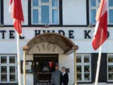 To personer står under en markise mærket 1902 ved indgangen til Hotel Hvide Kro med danske flag på hver side af døråbningen.