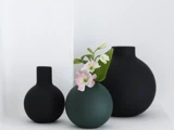 Tre runde, minimalistiske vaser – to i mat sort og én i mørkegrøn – står samlet. Den grønne vase indeholder små lyserøde og hvide blomster med grønne blade.