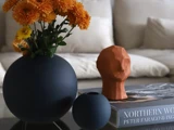 En rund vase med orange blomster, en lille vase, en geometrisk terrakotta-skulptur og en bog på et glasbord foran en beige sofa.