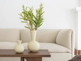 To cremefarvede vaser med grønne, bladrige grene står på et træbord foran en lysbeige sofa i en minimalistisk, moderne stue.