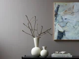 To hvide vaser med bare grene står på et mørkt bord ved siden af bøger og en skulptur, mens der hænger et abstrakt maleri på en grå væg.