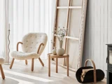 En hyggelig stol med fåreskind, et sidebord med blomstervaser, en brændeholder og et stort lærred ved siden af de hvide gardiner i et lyst, minimalistisk rum.