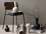 En moderne stol med bøger og vaser på og omkring den, et sammenrullet stykke papir og en gren i en vase, alt sammen placeret på et lyst trægulv foran en beige væg.