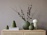 Minimalistisk indretning med grønne vaser, tørrede grene, en hvid abstrakt skulptur, to bøger og grønne træformede pyntegenstande på et træskab foran en taupefarvet væg.