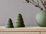 To grønne, træformede dekorationer står på en flettet overflade ved siden af en rund grøn vase med grene foran en grå væg.
