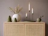 Minimalistisk indretning med en grøn lysestage, tændte stearinlys, en vase med tørret græs, to grønne juletræspynt og en lille rund vase på et kurveskab foran en taupefarvet væg.