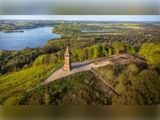 Luftfoto af et højt murstenstårn på en bakke omgivet af grønne træer, med en sø og et skovlandskab i baggrunden.