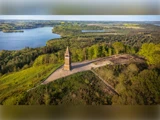 Luftfoto af et højt murstenstårn på en bakke omgivet af grønne træer, med en sø og et skovlandskab i baggrunden.
