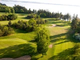Luftfoto af en frodig grøn golfbane med sandbunkere, træer og en sø i baggrunden i stærkt sollys.
