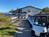 En række hvide golfvogne parkeret langs en asfalteret sti foran en toetagers bygning på en solrig dag.
