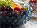 En struktureret glasskål fyldt med vindruer, kirsebær, jordbær og en appelsin står på en træplade med en anden frugtskål i baggrunden.