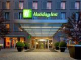 Indgangen til et Holiday Inn-hotel med et lysegrønt skilt, glasdøre, moderne baldakin og pottebuske langs gangstien.