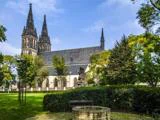 En historisk kirke med to spir, omgivet af grønne træer og en græsklædt park, med en stenbrønd og en bænk i forgrunden under en blå himmel.