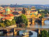 Farverigt bybillede af Prag med historiske bygninger, kupler, broer over Vltava-floden og en båd, der krydser i forgrunden under klar himmel.
