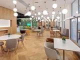 Moderne cafeteria med trægulv, runde og firkantede borde, forskelligfarvede stole, kugleformede hængelamper, potteplanter på bordene og store vinduer langs den ene side.
