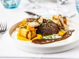 Et gourmetmåltid med stegt oksekød toppet med krydderurter, ristede kartofler, skiveskårne svampe, squashpuré og en brun sauce, serveret på en hvid tallerken med gaffel og kniv ved siden af.