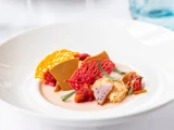 En gourmetdessert med farverige sukkerdekorationer, brune karamelsplinter, røde frugtstykker og fløde, kunstfærdigt anrettet på en hvid tallerken.
